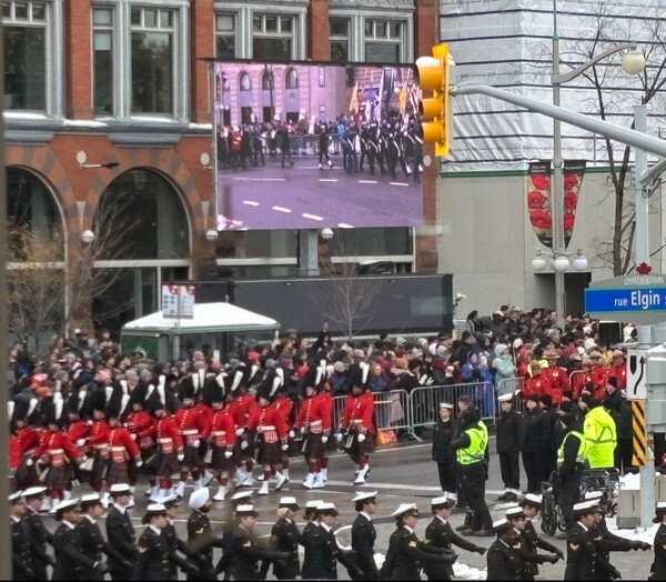 오타와 도심의 대형 스크린을 통해 리멤버런스 데이 중앙 추모식 현장이 생중계되고 있으며, 군인들이 엄숙하게 자리를 지키는 가운데 시민들이 추모 행렬을 지켜보고 있다.